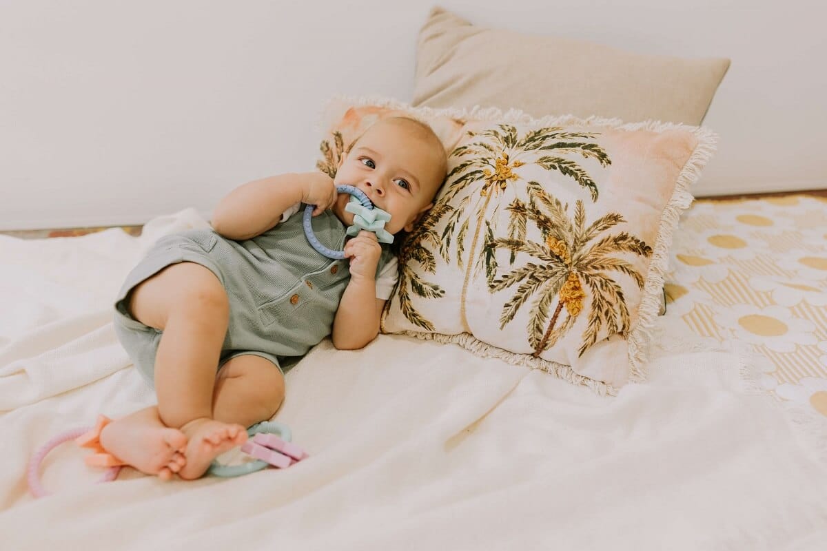 Baby lying on bed using a colorful assortment of chew necklaces and teething toys, including the Sunshine Teether, Sensory Fidget Ball, Rainbow Stacker and Teether Toy, jChews Chocolate Bar, and jChews Smartphone Teether. These safe and engaging toys are designed to provide soothing relief for teething infants.