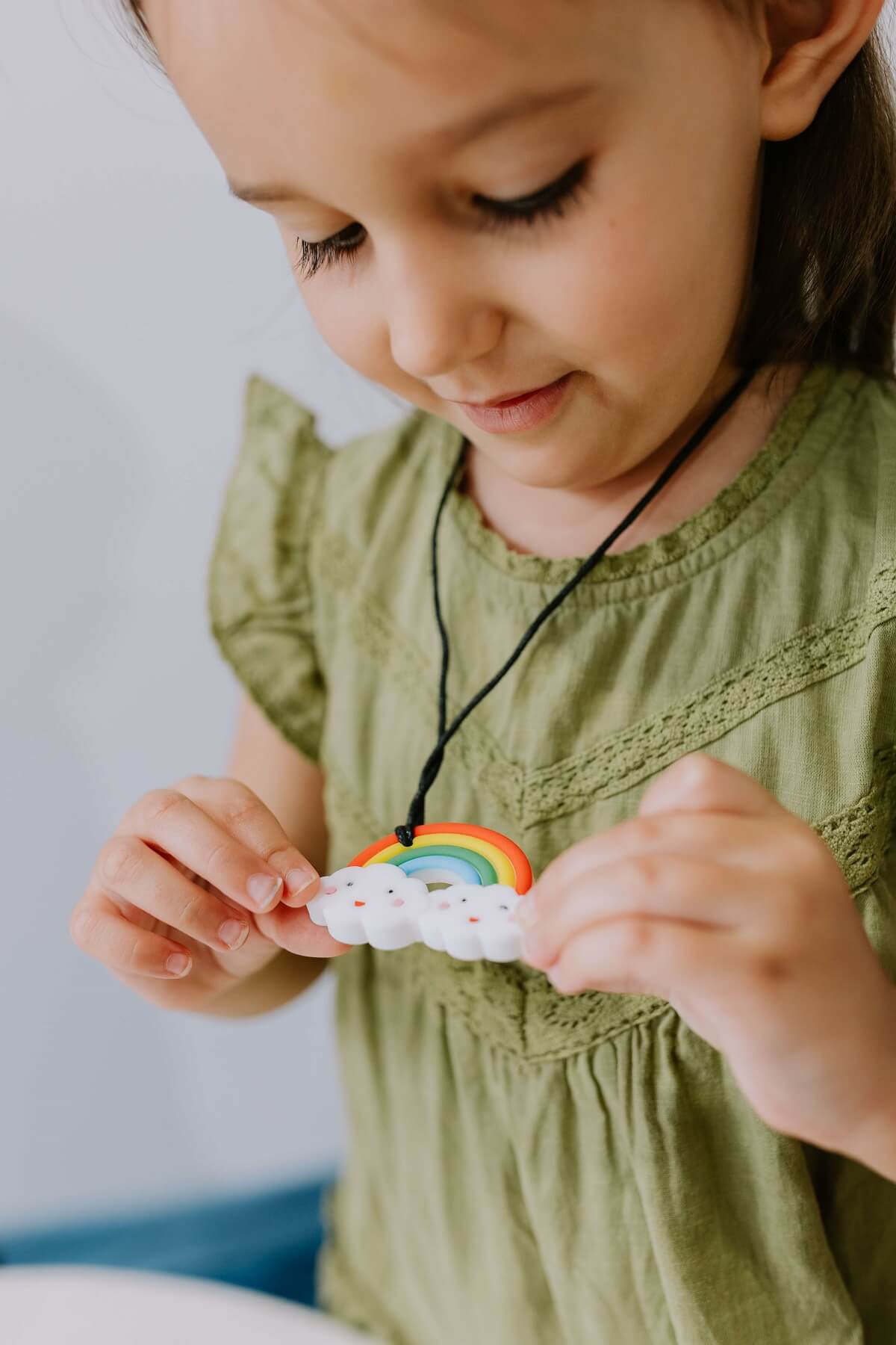 A close-up image of a child wearing a colorful silicone chew pendant necklace, designed to provide sensory stimulation and relief for teething discomfort. The pendant features a dinosaur shape with textured surfaces, and the child is happily chewing on it.